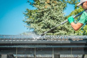 Person cleaning a rusted metal roof with a pressure washer on a sunny day, preparing the surface for painting. This image illustrates the step-by-step process of how to paint a rusted metal roof.