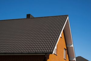 The image shows a close-up of a house with a metal roof that appears well-maintained and rust-free, set against a clear blue sky. The roof features neat, dark-colored metal tiles, and the house exterior is constructed with orange bricks. The image highlights the clean and modern aesthetic of a metal roof, offering a visual guide for those looking to learn "How To Paint Rusted Metal Roof" effectively.