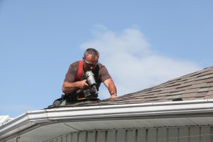 An experienced worker performing winter roof repairs on a house, using a nail gun to secure shingles on a sloped roof. The clear blue sky and the safety gear worn by the worker emphasize the importance of professional roof maintenance to prevent winter damage.