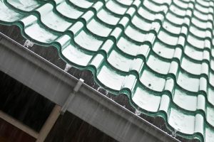 Close-up view of a green tiled roof under heavy rain, emphasizing the potential need for winter roof repairs. The image highlights the impact of severe weather conditions on roof integrity, underscoring the importance of regular maintenance and timely repairs to prevent damage during winter.