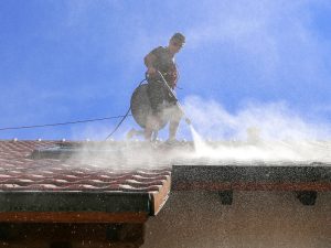 Technician performing roof restoration process using a pressure washer on a tiled roof under a clear blue sky.