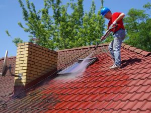 Worker pressure washing to remove moss on roof tiles near chimney and skylight for roof maintenance.